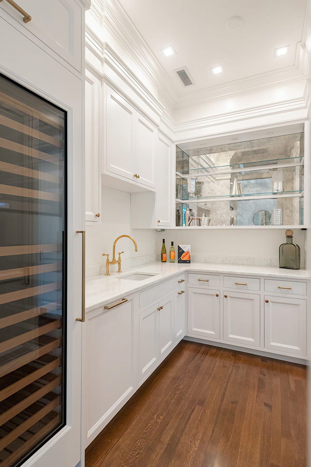 Butler’s pantry with white cabinetry, a wine refrigerator, brass fixtures, mirrored shelving, and wood flooring at the Chestnut Street Residence in Boston.